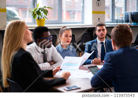 Multiethnic group of professionals engaged in a business meeting. African, Asian, and Caucasian team members discuss charts, collaborate on project in modern office setting. Multiethnic group of professionals engaged in a business meeting. African, Asian, and Caucasian team members discuss charts, collaborate on project in modern office setting. 113973543