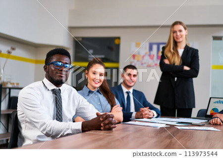 Multiethnic business team sits at meeting table in modern office. African, Caucasian, Arabic execs discuss strategy. Young woman leads diverse group. Colleagues with laptop, prepare presentation. Multiethnic business team sits at meeting table in modern office. African, Caucasian, Arabic execs discuss strategy. Young woman leads diverse group. Colleagues with laptop, prepare presentation. 113973544