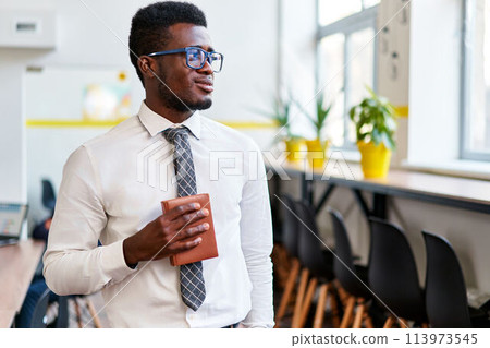 Confident African businessman in formal attire holds notebook in modern office space, looking away thoughtfully, reflecting corporate success and diverse workplace. Confident African businessman in formal attire holds notebook in modern office space, looking away thoughtfully, reflecting corporate success and diverse workplace. 113973545