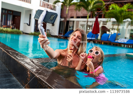 Mother and daughter enjoy ice cream while swimming in resort pool. Mom uses selfie stick for live stream video. Happy family vacation moment with tech interaction. Mom blogs, child smiles with treat. Mother and daughter enjoy ice cream while swimming in resort pool. Mom uses selfie stick for live stream video. Happy family vacation moment with tech interaction. Mom blogs, child smiles with treat. 113973586