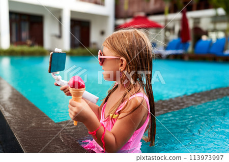 Girl in pink swimsuit enjoys ice cream, records with smartphone by poolside. Young influencer summer fun, live streaming pool time. Child engages followers with tasty treat cooling off in water. Girl in pink swimsuit enjoys ice cream, records with smartphone by poolside. Young influencer summer fun, live streaming pool time. Child engages followers with tasty treat cooling off in water. 113973997