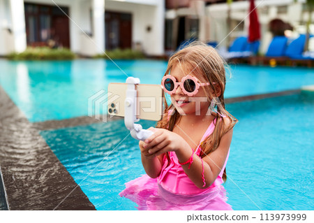 Smiling girl in pink swimwear records herself with phone on selfie stick at poolside. Child influencer enjoys ice cream, creates content for social media. Young vlogger at tropical resort. 113973999