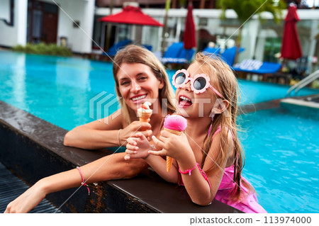 Smiling mom, daughter enjoy ice cream by poolside. Little girl in pink swimsuit, goggles eats dessert while her mother looks on happily. Family fun, summer leisure at tropical resort during vacation. Smiling mom, daughter enjoy ice cream by poolside. Little girl in pink swimsuit, goggles eats dessert while her mother looks on happily. Family fun, summer leisure at tropical resort during vacation. 113974000