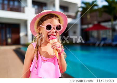Little girl in pink swimsuit, sunhat enjoys ice cream near pool. Child cools off with sweet treat on summer day. Bright sunshine, holiday relaxation, playful kid at resort. Cheerful moment by water. Little girl in pink swimsuit, sunhat enjoys ice cream near pool. Child cools off with sweet treat on summer day. Bright sunshine, holiday relaxation, playful kid at resort. Cheerful moment by water. 113974005