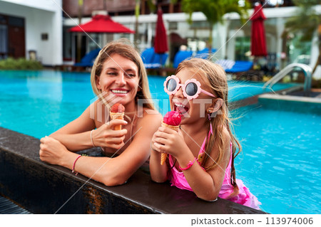 Smiling mom and daughter enjoy ice cream by poolside. Girl in pink swimsuit, flower sunglasses has fun licking cone. Happy family moment in summer. Vacation vibe at luxury resort with pool background. Smiling mom and daughter enjoy ice cream by poolside. Girl in pink swimsuit, flower sunglasses has fun licking cone. Happy family moment in summer. Vacation vibe at luxury resort with pool background. 113974006