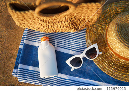 Sunscreen bottle on striped towel next to woven bag, straw hat, and white sunglasses on sandy beach suggests smart sun safety habits while soaking up the seaside ambience on a sunny day. 113974013
