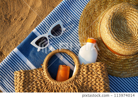 Summer accessories on sandy beach at sunset. Woven bag with sunscreen inside next to stylish sun hat. White sunglasses lie on blue striped towel. Vacation essentials for sun protection. Summer accessories on sandy beach at sunset. Woven bag with sunscreen inside next to stylish sun hat. White sunglasses lie on blue striped towel. Vacation essentials for sun protection. 113974014