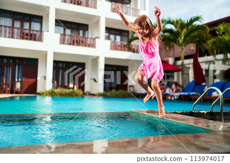 Happy girl in pink swimsuit jumps with joy into outdoor pool at sunny resort. Child enjoys summer vacation, fun splashing water. Excitement, leisure activity, swimming, playful moment captured. 113974017