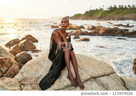 Queer black person in luxury dress, jewelry sits on rocks in ocean. Lgbtq ethnic fashion model wearing jewellery dressed in posh gown poses gracefully in tropical seaside location. Pride LGBTQIA 113974026