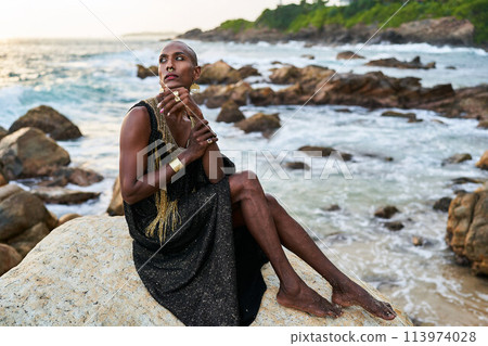 Non-binary black person in luxury dress, golden jewelry on beach rocks in ocean. Trans ethnic fashion model wearing jewellery in posh gown poses gracefully in tropical seaside location on sunset. 113974028