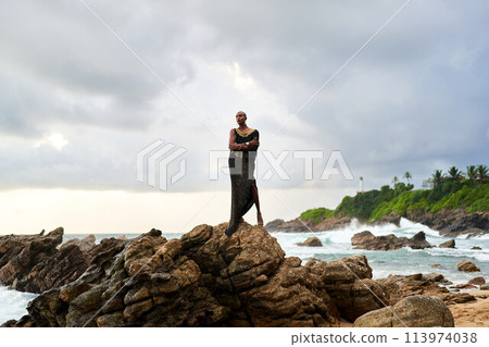 Non-binary black godlike person poses gracefully standing on rocks in ocean. Trans ethnic fashion model in posh dress and jewellery on rocky beach by storm . Lgbtq. Pride month 113974038