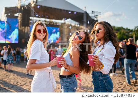 Group of smiling young women with sunglasses enjoy summer music festival on beach holding colorful drinks, casual fashion, sunset party vibe with concert stage in background and crowd gathering. 113974039