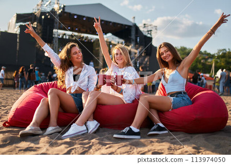 happy girls sit on red bean bag chairs at beach music festival, raise arms, hold drinks. Teen friends enjoy summer concert, casual clothes, sneakers style. Outdoor event, fans relax, vibe together. 113974050
