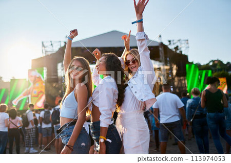 Group of girls enjoy summer music festival on sandy beach at sunset. Friends dance, celebrate near stage, vibrant colors. Casual fashion, happy youth lifestyle, outdoor concert event. Group of girls enjoy summer music festival on sandy beach at sunset. Friends dance, celebrate near stage, vibrant colors. Casual fashion, happy youth lifestyle, outdoor concert event. 113974053