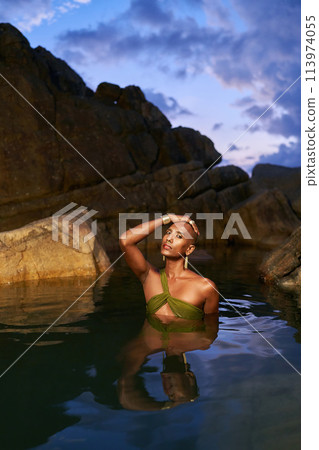 Gender fluid black person dives out from water in natural still water pool. Queer ethnic fashion model in long revealing clothes poses gracefully waist deep in middle of crystal clear lake at night. Gender fluid black person dives out from water in natural still water pool. Queer ethnic fashion model in long revealing clothes poses gracefully waist deep in middle of crystal clear lake at night. 113974055