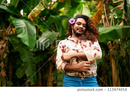 South Asian gay man with floral shirt, blue jeans stands in tropical foliage, purple flower in hair. Confident smile, celebrates inclusivity, pride, LGBT identity amidst natures embrace. 113974058