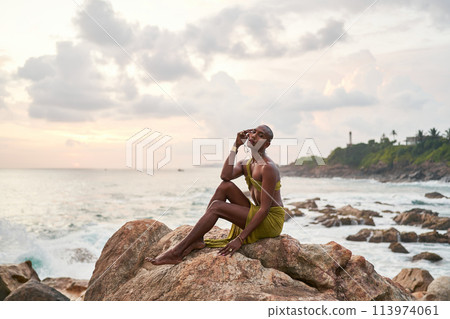 Gay bipoc graceful man poses sitting high on rocks in ocean at sunset. Homosexual slim ethnic fashion model in tropical maxi dress on top of rocky hill above storm at dusk. Pride LGBTQIA Gay bipoc graceful man poses sitting high on rocks in ocean at sunset. Homosexual slim ethnic fashion model in tropical maxi dress on top of rocky hill above storm at dusk. Pride LGBTQIA 113974061