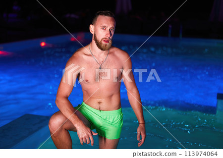 Seductive homosexual man celebrating gay pride with at night LGBTQ pool party in a private villa swimming pool. Bisexual male in swimwear posing on camera. 113974064