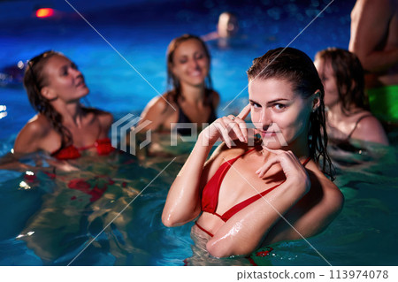 Pretty woman in red bikini hanging out with friends at night pool party in beach club. Nice girl in hot swimwear enjoy summer. People have fun swimming at water in luxury private villa swimming pool. 113974078