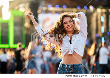 Happy young woman dances at sunset outdoor music fest on beach. Carefree girl enjoys summer concert, waves hands, smiles in crowd. Vibrant youth lifestyle celebrated at festival event by sea. 113974079