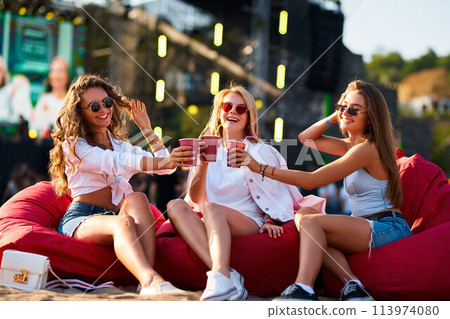 Girls in summer outfits sit on red bean bag chairs, clinking cups at beach music festival. Laugh, enjoying fest vibes, relax with drinks, sunset, ocean backdrop. Casual celebration cheerful friends. Girls in summer outfits sit on red bean bag chairs, clinking cups at beach music festival. Laugh, enjoying fest vibes, relax with drinks, sunset, ocean backdrop. Casual celebration cheerful friends. 113974080