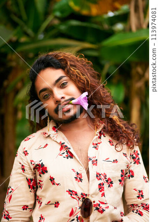 Gay man with curly hair and floral shirt stands in garden, purple flower in mouth. Expresses freedom, LGBT pride, gender fluidity. Relaxed, confident pose in natural setting. Symbolizes love, spring. 113974081