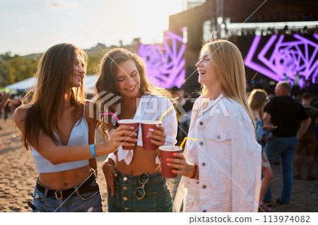 Women clink cups at beach music fest, happy friends toast with drinks in sunshine. Outdoor party vibe, casual summer fun with crowd and festival stage backdrop. Cheerful group celebrate, laughter. Women clink cups at beach music fest, happy friends toast with drinks in sunshine. Outdoor party vibe, casual summer fun with crowd and festival stage backdrop. Cheerful group celebrate, laughter. 113974082