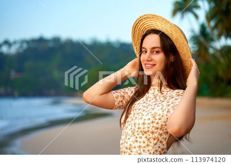 Smiling young woman in straw hat on tropical beach at sunset. Relaxed tourist enjoys warm evening by sea. Leisure in nature, summer travel, vacation mood. Vibrant ocean backdrop with palm trees. 113974120