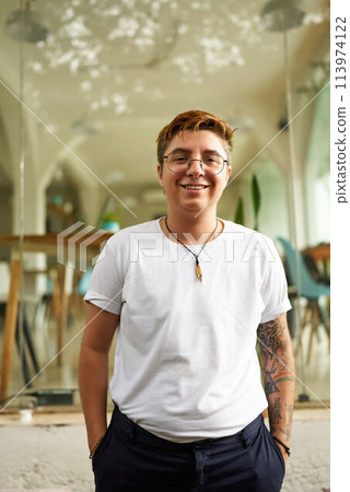 Gen Z Transgender pro in smart casual attire stands confident in modern office environment. Young, inclusive, LGBTQ friendly, glasses, smiling, represents workplace diversity, gender identity. 113974122