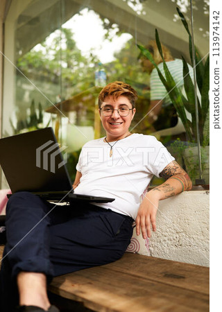 Transgender Gen Z Freelancer works remotely on laptop in cozy LGBT-friendly coworking space. Smiling, tattooed individual in casual clothes enjoys flexibility, achieves work-life balance. Transgender Gen Z Freelancer works remotely on laptop in cozy LGBT-friendly coworking space. Smiling, tattooed individual in casual clothes enjoys flexibility, achieves work-life balance. 113974142