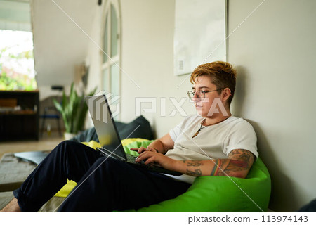 Transgender pro works on laptop from cozy bean bag in modern coworking space. Focused on screen, person enjoys flexible working environment. Inclusive workplace, remote job and casual office setup. 113974143