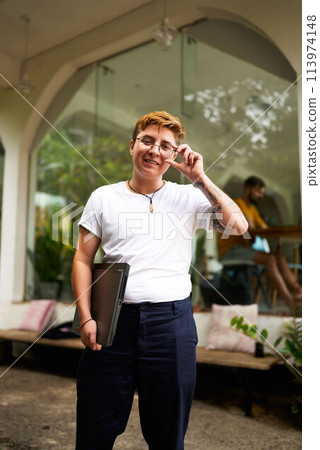 Transgender professional stands confidently in an office with laptop. Smiling, glasses-adjusting pose reflects inclusive, modern work environment. Casual style, tech in hand. Transgender professional stands confidently in an office with laptop. Smiling, glasses-adjusting pose reflects inclusive, modern work environment. Casual style, tech in hand. 113974148