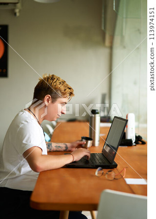 Transgender freelancer works on laptop in coworking space, sips tea from vacuum flask mug. Casual pro in creative office setting engages in digital tasks, enjoys beverage, reflects agile work culture. 113974151