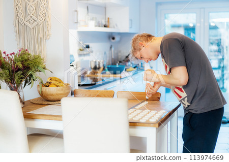Concentrated young man placing batter on silicone baking sheet to make French macaroons dessert in light modern kitchen at home. Preparing steps for baking cookies. Culinary hobby. Selective focus 113974669