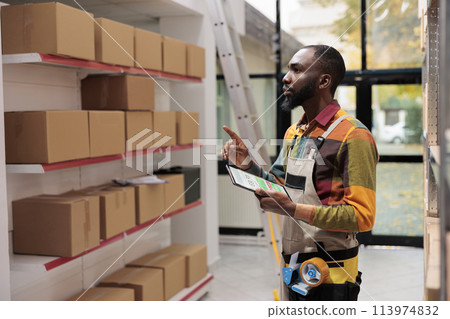 Storehouse employee using inventory app on digital tablet for goods management. African american warehouse manager wearing protective industrial overall analyzing online checklist Storehouse employee using inventory app on digital tablet for goods management. African american warehouse manager wearing protective industrial overall analyzing online checklist 113974832