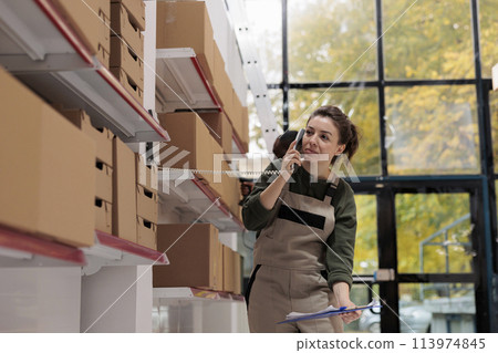 Storage room worker discussing inventory details with remote manager at landline phone during work shift in warehouse. Employee working at client orders, preparing products for shipping 113974845