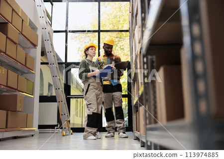 Diverse team wearing helmet and industrial overall, checking cardboard boxes for packages shipping in warehouse. Storage room employess preparing customers orders while doing goods quality control 113974885