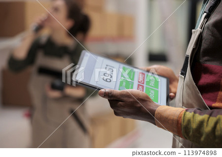Supervisor checking inventory report using tablet computer, working at clients order preparing delivery in warehouse. African american employee supervising boxes in mail sorting center. Close up 113974898