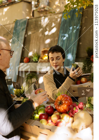 Female vendor arranging organic produce on greenmarket stand while assisting elderly male client with purchasing fresh fruits and vegetables. Senior man buying locally grown bio food products. Female vendor arranging organic produce on greenmarket stand while assisting elderly male client with purchasing fresh fruits and vegetables. Senior man buying locally grown bio food products. 113975028