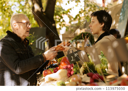 Young vendor giving freshly harvested organic carrots at farmers market stall booth. Elderly customer buying locally grown vegetables from female merchant at greenmarket stand. 113975030