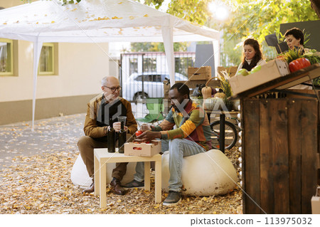 African american customer sitting at a farmers market table with a local vendor, having homemade wine. Black male client visiting an organic autumn festival and enjoying wine and food tasting. 113975032
