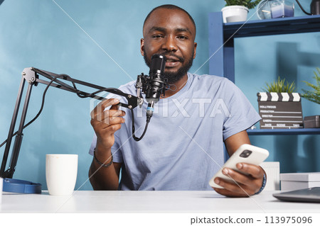 Vlogger creating podcast content while reading news from smartphone and looking at camera. African american man holding mobile phone and answering question while streaming live 113975096