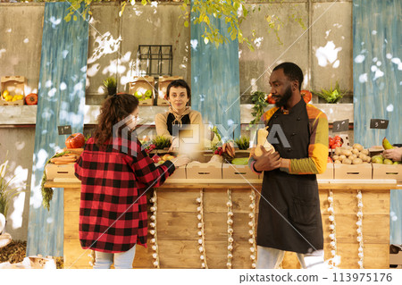 Black male farmer showing natural organic seasonal produce to customer at greenmarket kiosk. Group of vendors and clients at harvest fair festival, browsing freshly harvested fruits and vegetables. 113975176