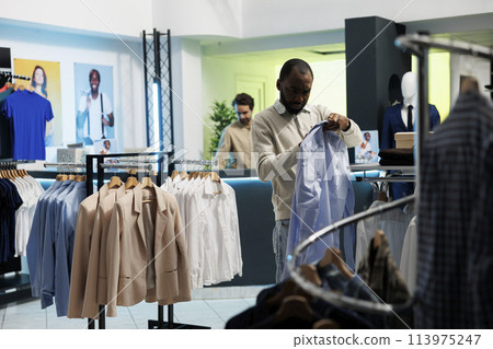 African american buyer checking size on shirt label while choosing trendy formal wear in shopping mall boutique. Young man browsing fashionable apparel on hanger in showroom African american buyer checking size on shirt label while choosing trendy formal wear in shopping mall boutique. Young man browsing fashionable apparel on hanger in showroom 113975247