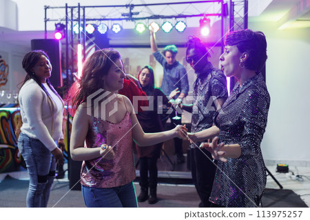 Carefree women dancing in crowded nightclub at live music concert. Cheerful young girlfriends partying with friends and dancing at clubbers gathering event during dj performance 113975257