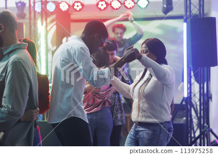 Young man and woman friends holding hands while dancing at nightclub party. African american couple clubbing and moving on dancefloor while attending discotheque gathering event 113975258