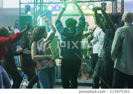 Active people dancing and raising hands while partying in nightclub. Young men and women moving to electronic music beats and relaxing on crowded dancefloor at discotheque 113975261