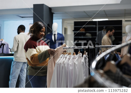 Smiling african american woman holding jacket on hanger while shopping for outfit in clothing store. Fashion boutique customer exploring apparel rack and selecting formal wear 113975282