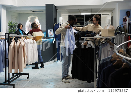 Clothing store customer trying on shirt and getting advice from boutique employee. African american man holding apparel to body while choosing size with fashion outlet assistant 113975287