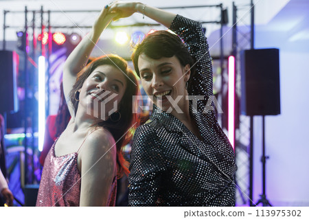 Cheerful girlfriends improvising moves on dancefloor, celebrating and partying in nightclub. Young carefree women friends dancing and having fun at discotheque event in club 113975302
